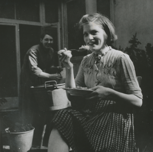 876777 Portret van een jongedame die voedsel eet van een bord, op de buitenplaats bij de gaarkeuken die ingericht is in ...